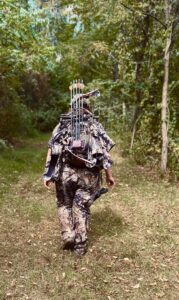 Woman walking into the woods with her archery gear strapped to her back.
